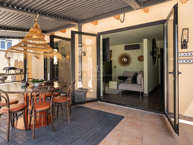 Une terrasse couverte avec une table à manger ronde, une suspension en rotin et des baies vitrées ouvrant sur le salon.