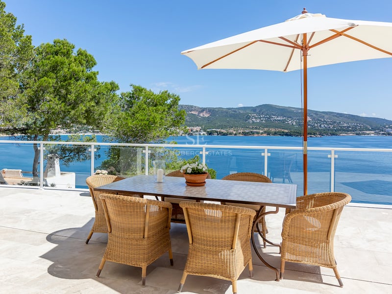 Espace repas extérieur avec chaises en osier et parasol blanc sur terrasse en pierre surplombant mer et montagnes.