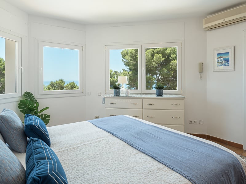 Chambre blanche lumineuse avec de grandes fenêtres vue mer, literie bleue et porte vers un balcon ensoleillé.