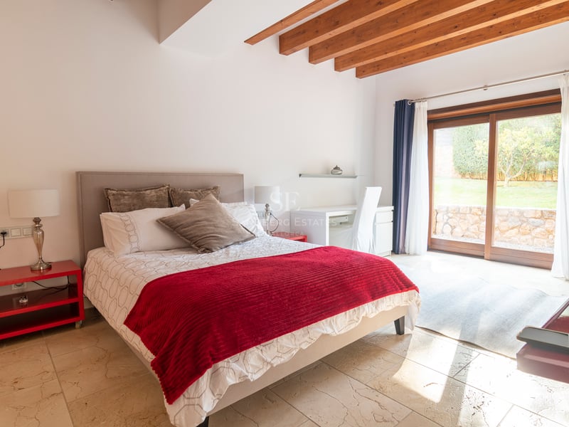 Chambre lumineuse avec accents rouges, sol en pierre, poutres en bois et baies vitrées donnant sur le jardin.