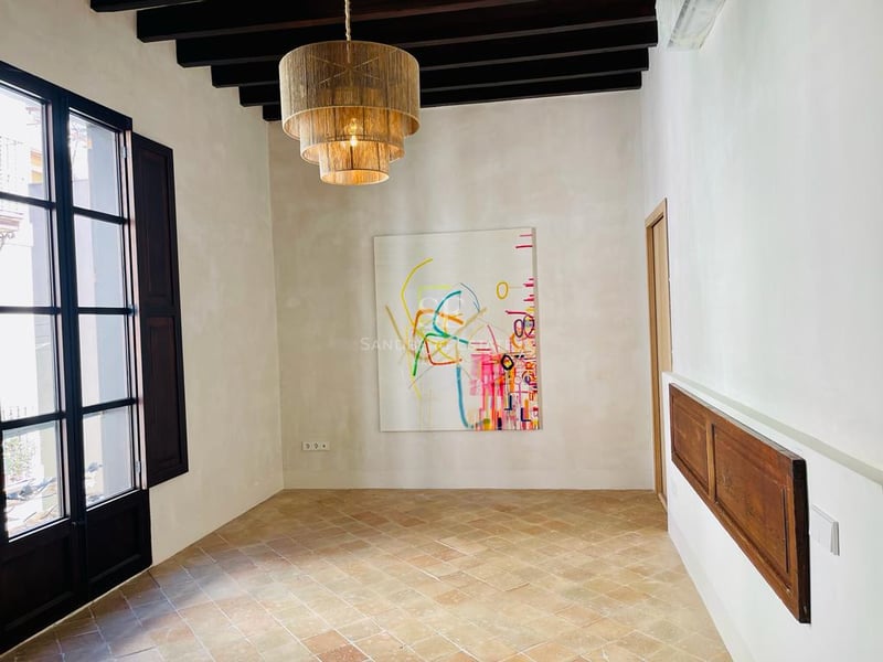 Pièce avec sol en terre cuite, poutres en bois sombre, grande fenêtre noire et lustre en rotin avec art moderne.