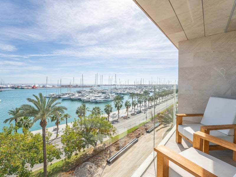 Balcon moderne avec chaises en bois et garde-corps en verre donnant sur un port de plaisance et des palmiers.