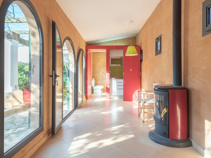 Pièce ensoleillée avec murs en terre cuite, portes vitrées cintrées, poêle à bois rouge et vue sur le jardin.