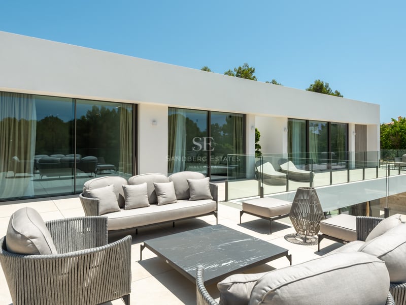 Terrasse de villa moderne avec salon de jardin gris, garde-corps en verre et larges baies vitrées.