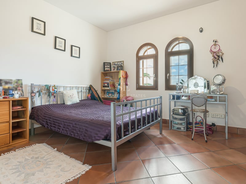 Chambre avec sol en terre cuite, lit en métal, fenêtres cintrées en bois et placards intégrés sous lumière naturelle.