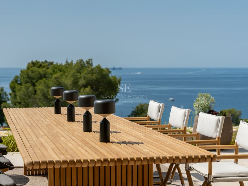 Table à manger moderne en bois avec lampes design sur une terrasse ensoleillée surplombant la Méditerranée.
