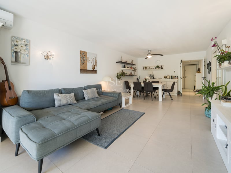 Salon lumineux avec un grand canapé en L bleu, des meubles blancs, un coin repas et un sol carrelé.