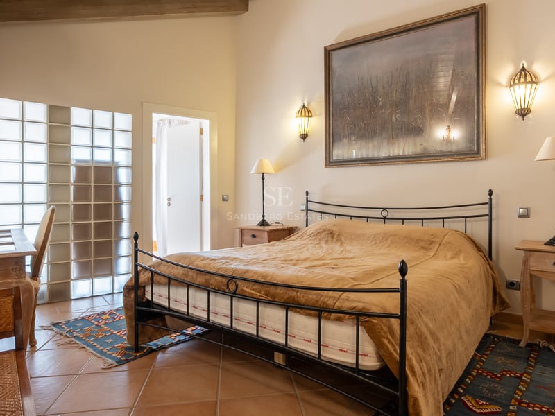 Chambre avec lit en fer forgé, sol en terre cuite et mur en briques de verre sous des poutres apparentes.