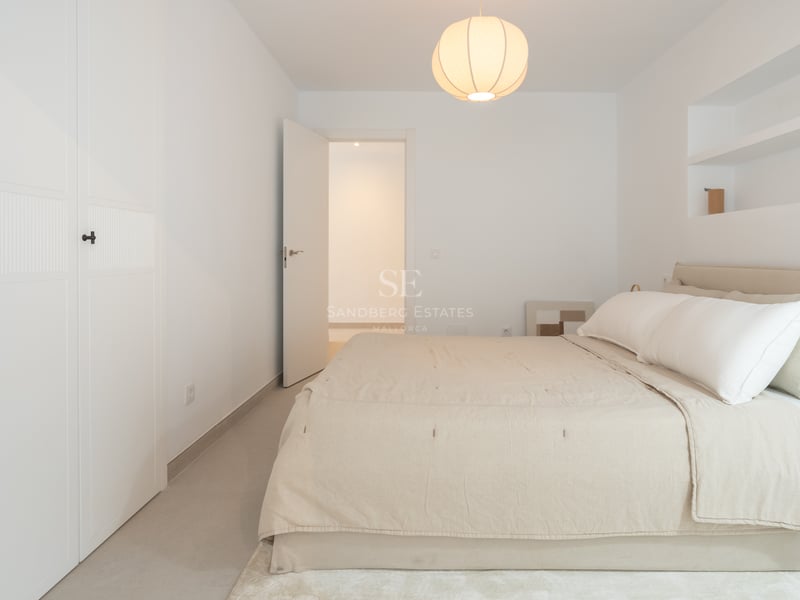 Une chambre minimaliste lumineuse avec un grand lit, du linge blanc, une suspension ronde et une porte blanche ouverte.