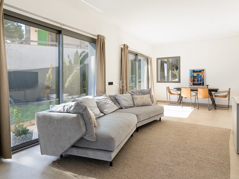 Salon lumineux avec grand canapé gris, baies vitrées donnant sur le jardin et coin repas avec chaises en cuir.