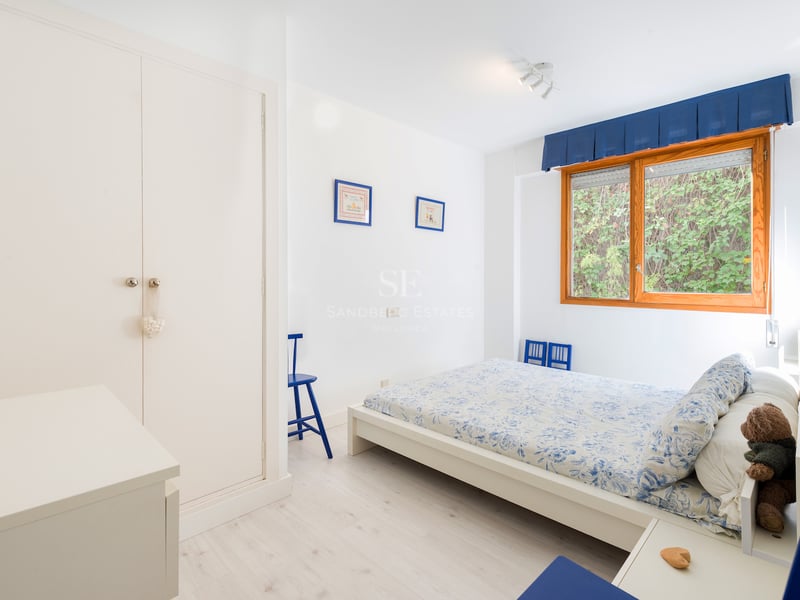Chambre blanche et lumineuse avec une fenêtre à cadre en bois, un placard intégré et une literie florale bleue.