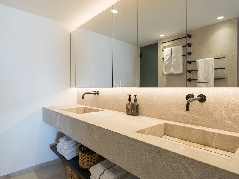Salle de bain de luxe avec double vasque intégrée en pierre, miroirs rétroéclairés et robinetterie noire mate.