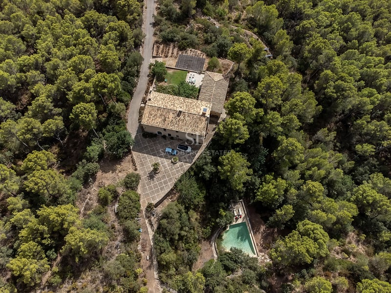 Vue aérienne d'une maison en pierre avec toit en terre cuite, cour et piscine, entourée d'une forêt de pins dense.