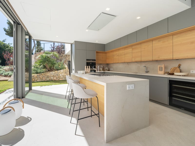 Cuisine moderne grise et bois avec îlot et taburetes, s'ouvrant sur un jardin ensoleillé par de grandes baies vitrées.