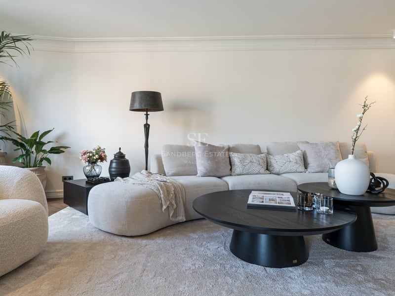 Modern living room with a curved beige sofa, black round coffee tables, and a plush rug in a bright neutral room.