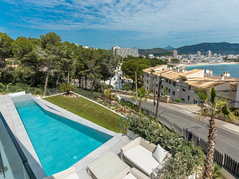 Une longue piscine turquoise sur une terrasse avec garde-corps en verre, surplombant une ville côtière et des montagnes.