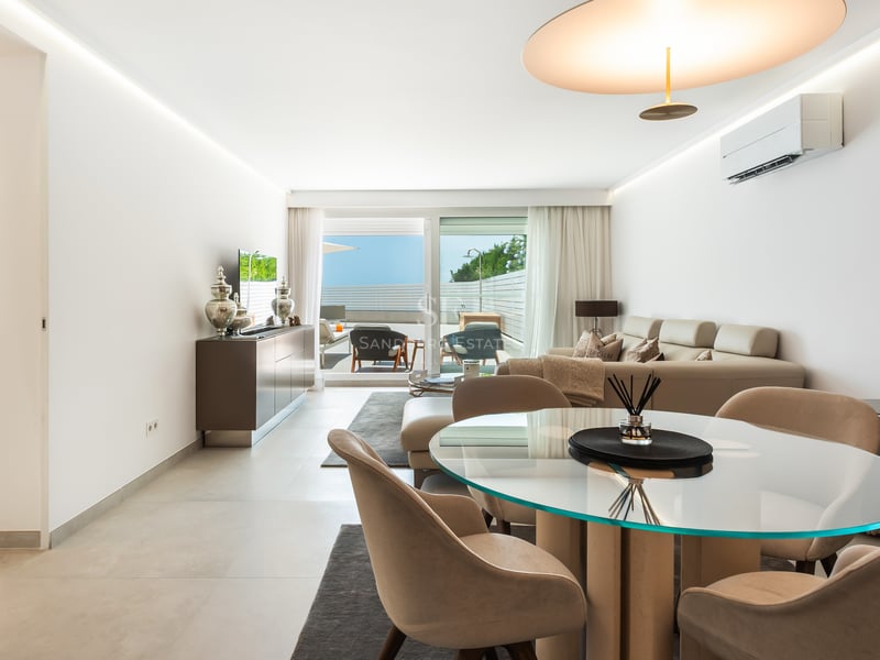 Salon lumineux avec table à manger ronde en verre, canapé crème et accès à une terrasse ensoleillée.