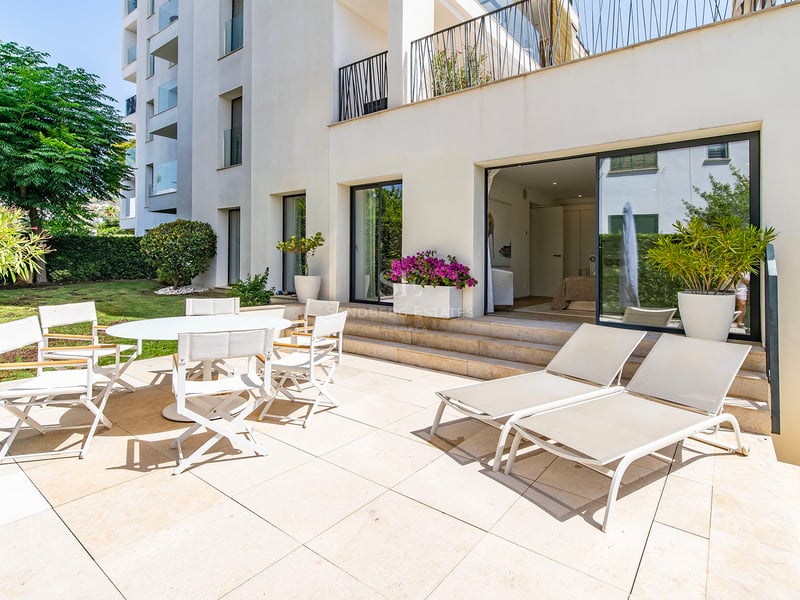 Terrasse ensoleillée avec coin repas blanc et chaises longues à côté d'un jardin et d'un immeuble moderne.