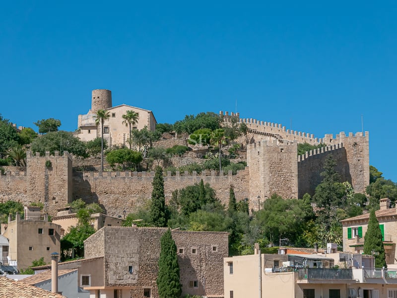 Stone walls and towers of a medieval fortress on a hill under a blue sky in a Mallorcan village.