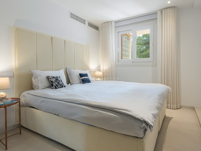 Chambre lumineuse avec tête de lit capitonnée crème, draps blancs et lampes de chevet modernes près d'une fenêtre.
