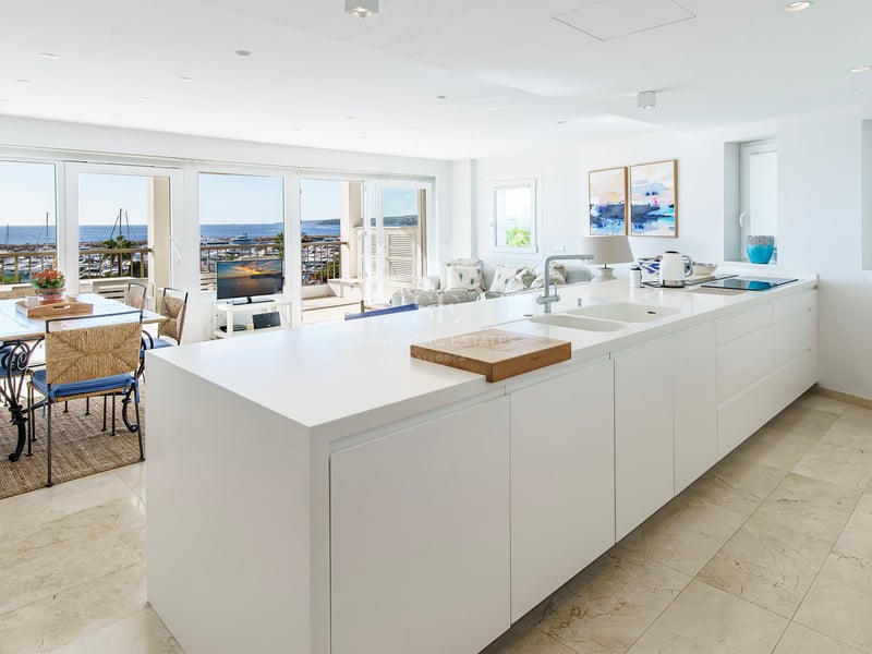 Ilot de cuisine blanc minimaliste s'ouvrant sur une salle à manger avec vue panoramique sur le port et la mer.