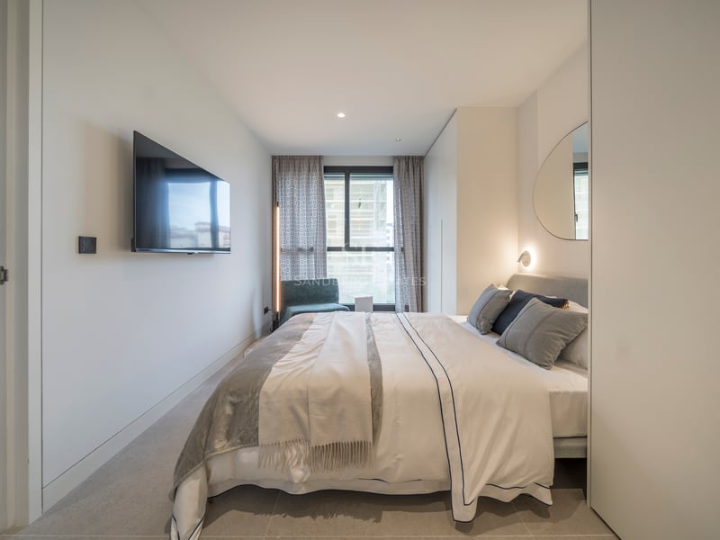 Chambre lumineuse avec TV murale, lit tapissé gris, miroir organique et grande fenêtre.