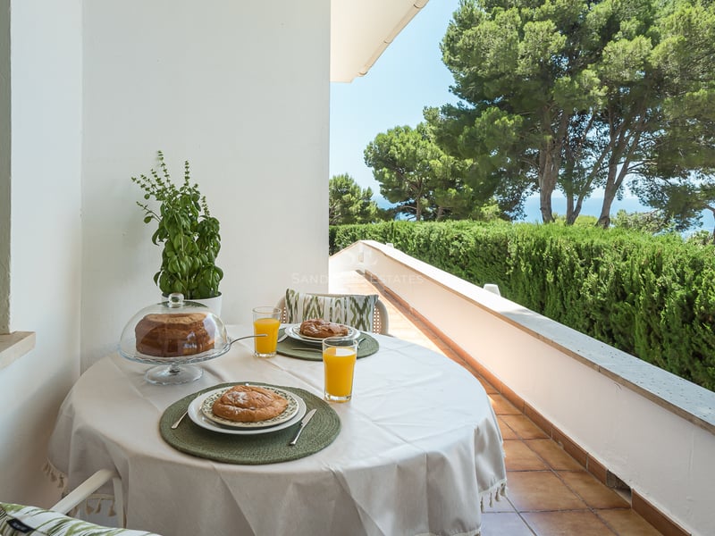 Balcon blanc avec table de petit-déjeuner dressée, surplombant des pins et la mer bleue au loin.