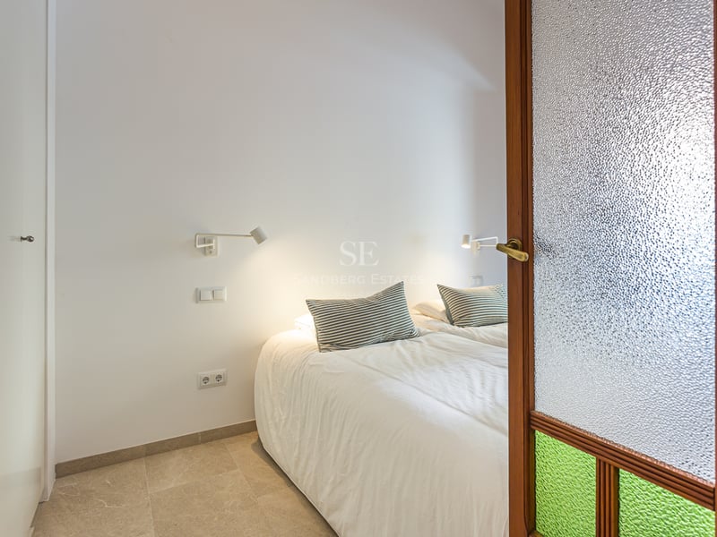 Vue d'une chambre moderne à travers une porte en bois avec verre texturé, lit blanc et liseuses murales.