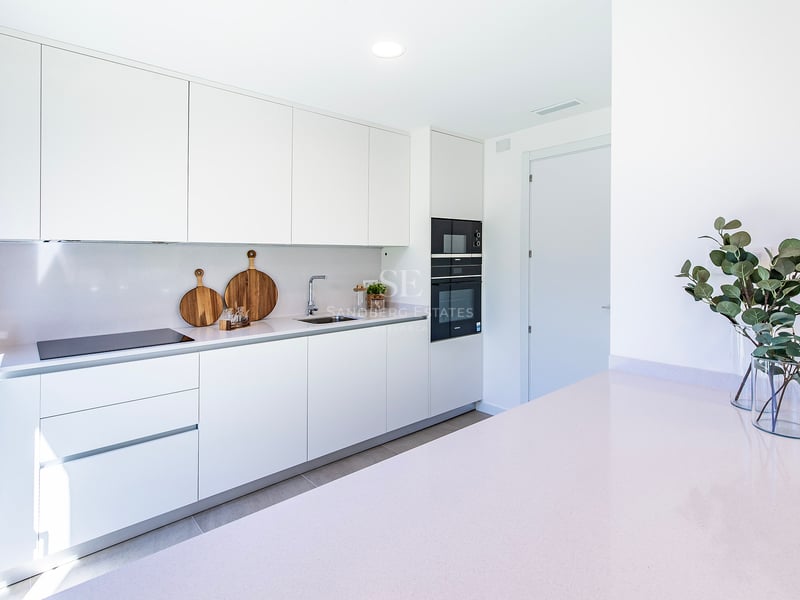 Ljust vitt kök med minimalistiska skåp, integrerade vitvaror och bänkskiva i ljus sten.