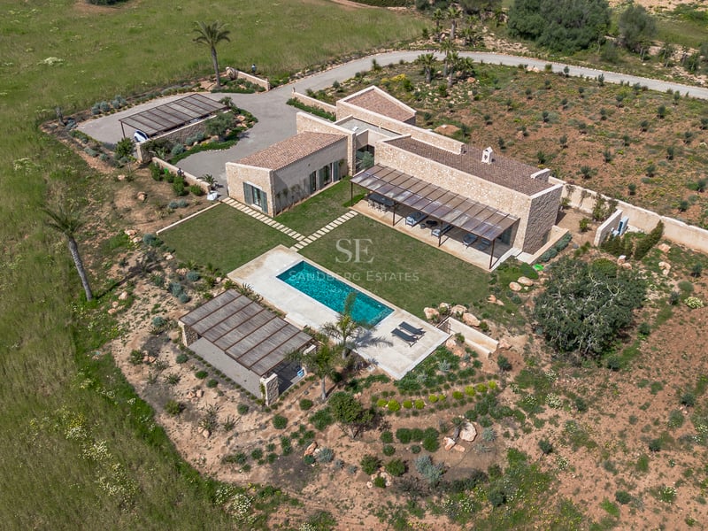 Vista aérea de una villa moderna de piedra con piscina turquesa, jardín de césped y entorno natural en Mallorca.