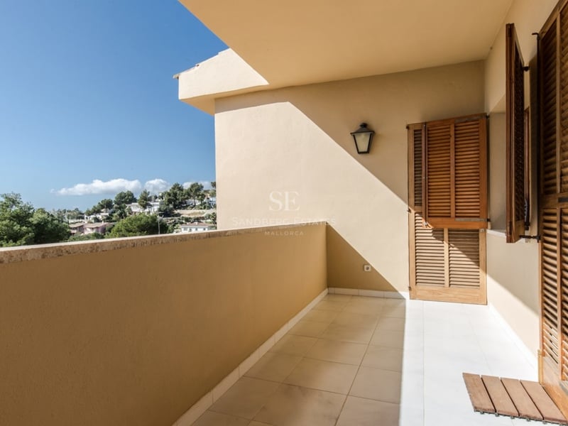 Balcon couvert avec volets en bois et carrelage offrant une vue sur la mer et la verdure sous un ciel bleu.