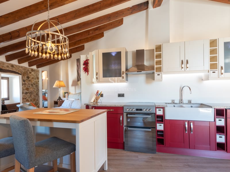 Cuisine avec placards rouges, poutres en bois apparentes et mur en pierre naturelle avec îlot central.