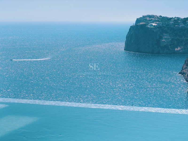 Bord d'une piscine à débordement dominant une mer bleue avec des falaises côtières et un sillage de bateau.