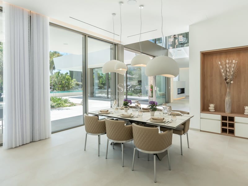 Salle à manger moderne avec grande table, chaises en cuir beige et baies vitrées donnant sur une piscine.