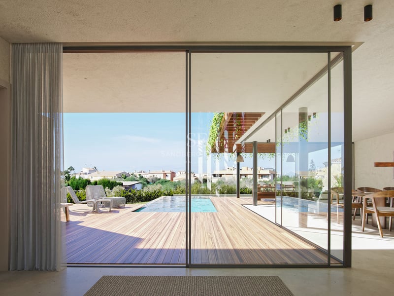 Vue depuis le salon à travers de grandes baies vitrées vers une terrasse en bois avec piscine et chaises longues.