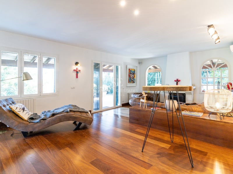 Salon lumineux avec parquet, chaise longue en cuir, canapé marron et cheminée blanche près de grandes fenêtres.