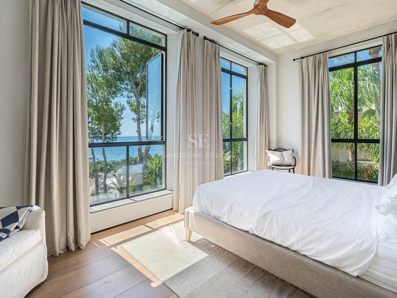 Chambre lumineuse avec lit blanc, parquet, ventilateur de plafond et grandes fenêtres offrant une vue sur la mer.