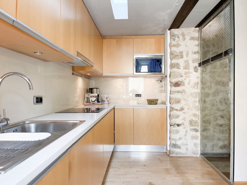 Cuisine moderne avec placards en bois clair, plans de travail blancs et mur en pierre apparente.