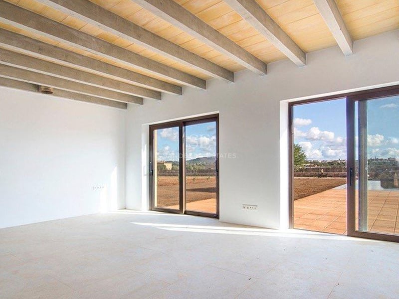 Grande pièce vide avec poutres apparentes en béton et bois, murs blancs et baies vitrées coulissantes.