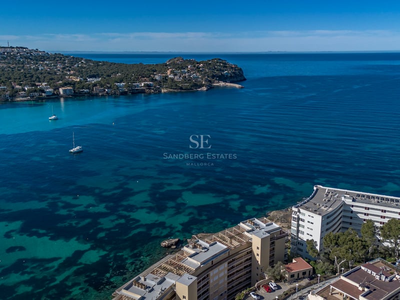 Vue aérienne d'une baie turquoise avec des voiliers à l'ancre et des bâtiments côtiers sous un ciel bleu.