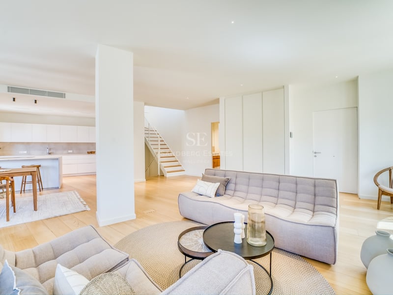Modern open-plan living room featuring tufted grey sofas, light oak flooring, and a kitchen in the background.
