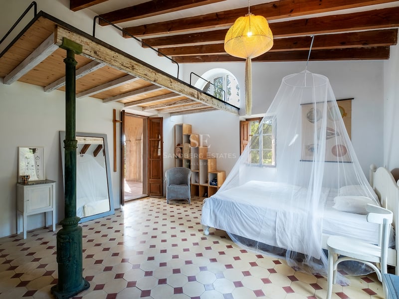 Grande chambre avec lit blanc, moustiquaire, mezzanine en bois, poutres apparentes et sol en carreaux décoratifs.
