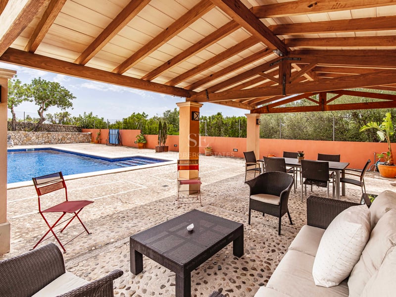 Terrasse couverte avec pergola en bois, sol en pierre naturelle, coin repas et salon face à une piscine bleue.