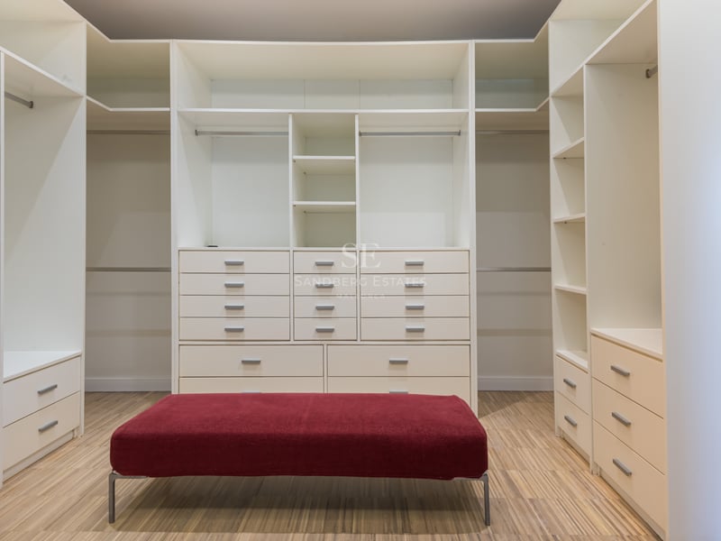 Dressing blanc spacieux avec de nombreuses étagères, tiroirs, sol en bois et un pouf en velours rouge au centre.