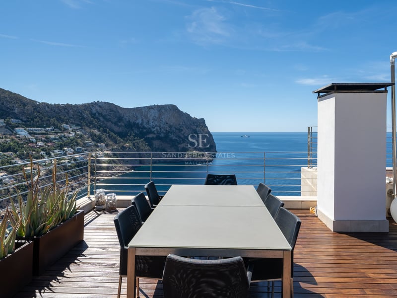 Terrasse moderne sur le toit avec une grande table à manger surplombant la mer et la côte escarpée sous un ciel bleu.