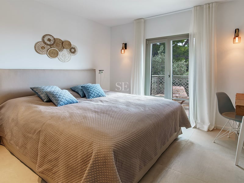Chambre moderne avec grand lit, literie beige, coussins bleus, bureau en bois et porte vitrée donnant sur un balcon.