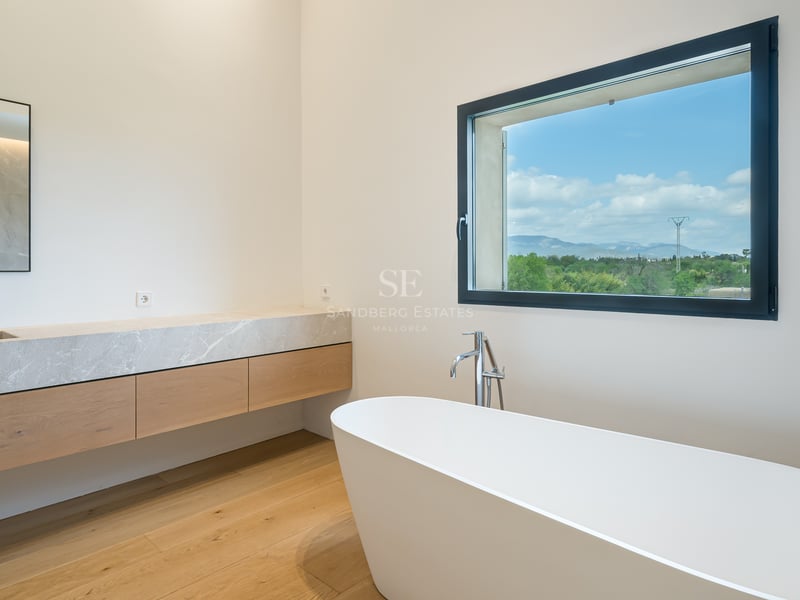 Salle de bains minimaliste avec baignoire îlot, meuble en chêne et vue sur les montagnes verdoyantes.