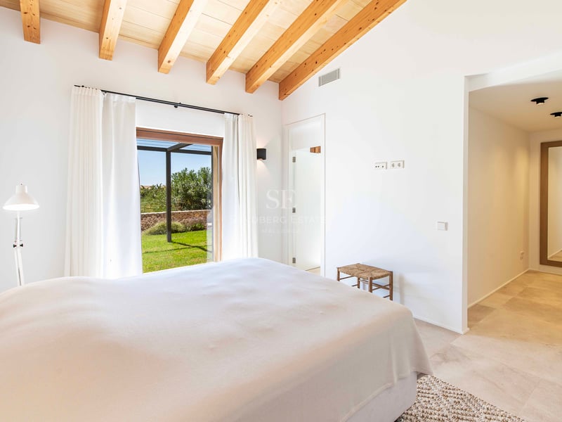 Chambre lumineuse avec poutres en bois, sol en pierre et accès direct à un jardin verdoyant par une porte vitrée.