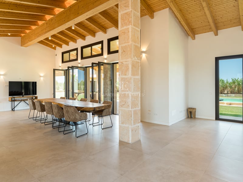 Amplio comedor con gran mesa de madera, columna de piedra natural, vigas de madera y acceso al jardín.