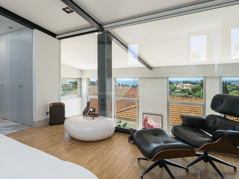 Chambre moderne avec fauteuil Eames, parquet en chêne et parois vitrées offrant une vue sur la mer et la ville.